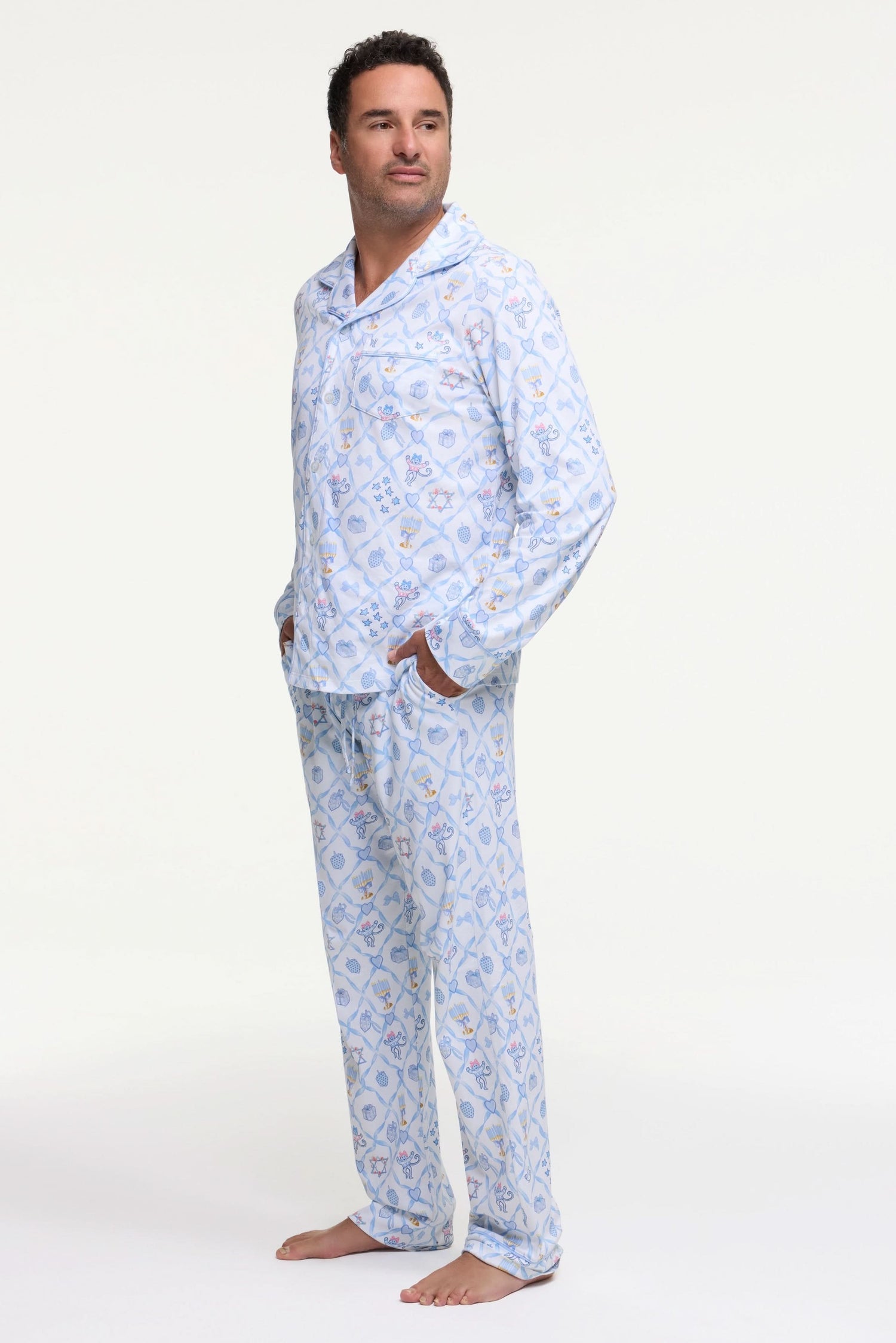 A man stands barefoot on a white background wearing Roller Rabbit's Mens Starry Nights Lattice Long Sleeve Polo Pajamas, featuring light blue and white patterns. He smiles with one hand in his pocket, highlighting the comfort of these matching PJs.
