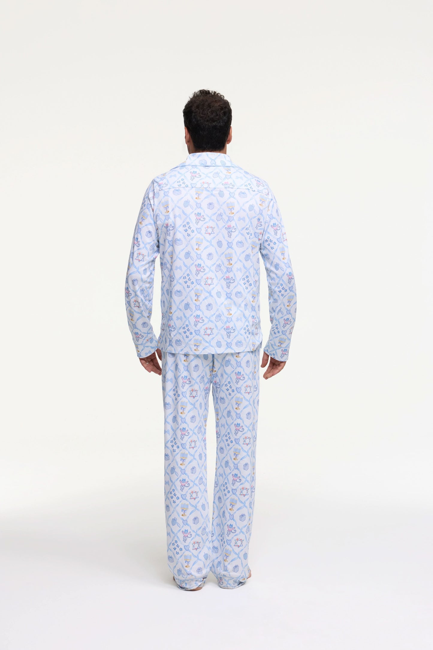 A man stands barefoot on a white background wearing Roller Rabbit's Mens Starry Nights Lattice Long Sleeve Polo Pajamas, featuring light blue and white patterns. He smiles with one hand in his pocket, highlighting the comfort of these matching PJs.