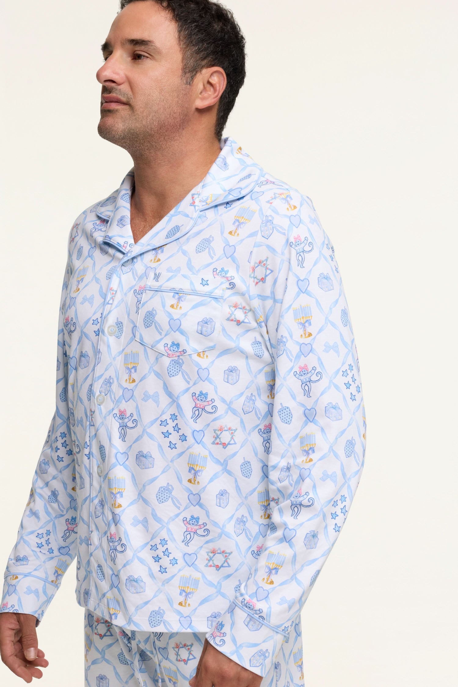 A man stands barefoot on a white background wearing Roller Rabbit's Mens Starry Nights Lattice Long Sleeve Polo Pajamas, featuring light blue and white patterns. He smiles with one hand in his pocket, highlighting the comfort of these matching PJs.