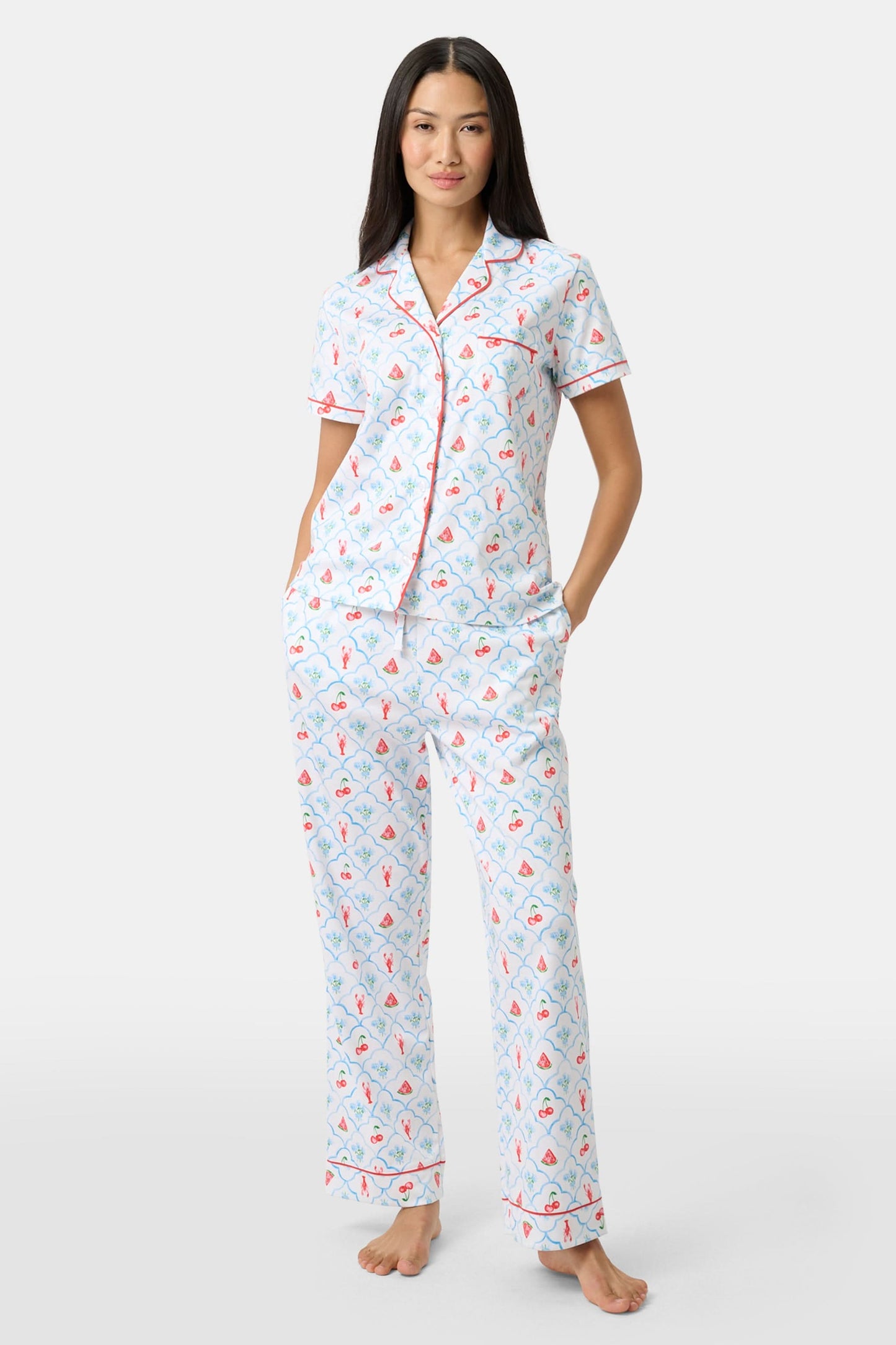 A woman in Roller Rabbit’s 4th of Jemina Fiora Pajamas and pink slippers relaxes on the bow of a blue boat at a marina while another person in white sleepwear stands nearby, partially out of frame.