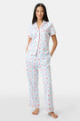 A woman in Roller Rabbit’s 4th of Jemina Fiora Pajamas and pink slippers relaxes on the bow of a blue boat at a marina while another person in white sleepwear stands nearby, partially out of frame.