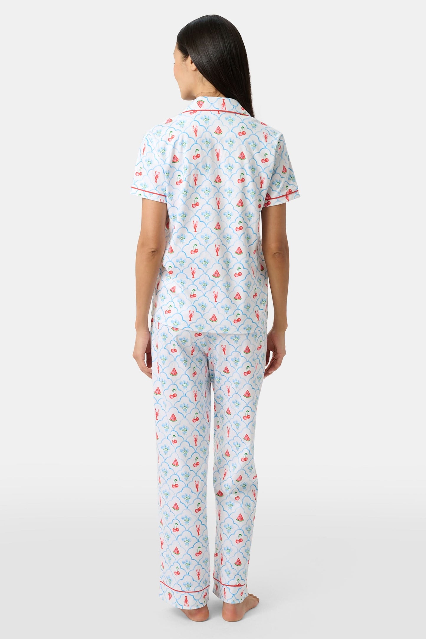 A woman in Roller Rabbit’s 4th of Jemina Fiora Pajamas and pink slippers relaxes on the bow of a blue boat at a marina while another person in white sleepwear stands nearby, partially out of frame.