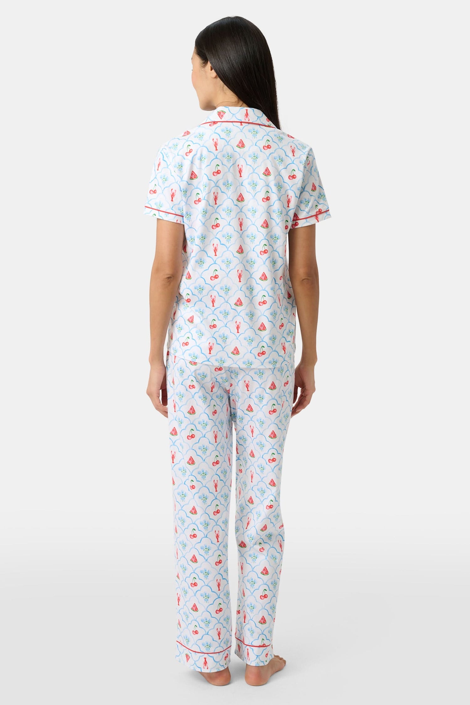 A woman in Roller Rabbit’s 4th of Jemina Fiora Pajamas and pink slippers relaxes on the bow of a blue boat at a marina while another person in white sleepwear stands nearby, partially out of frame.