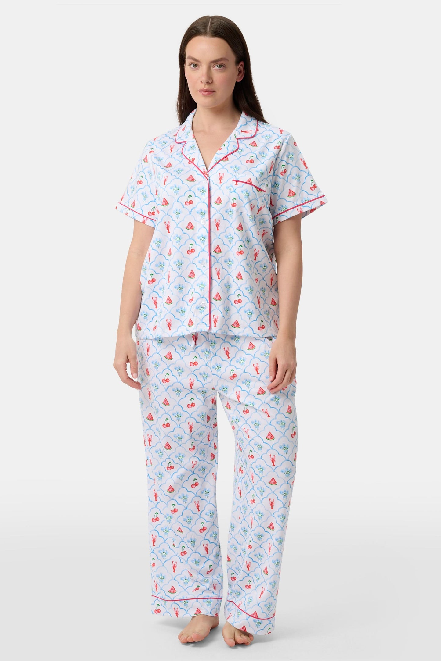 A woman in Roller Rabbit’s 4th of Jemina Fiora Pajamas and pink slippers relaxes on the bow of a blue boat at a marina while another person in white sleepwear stands nearby, partially out of frame.