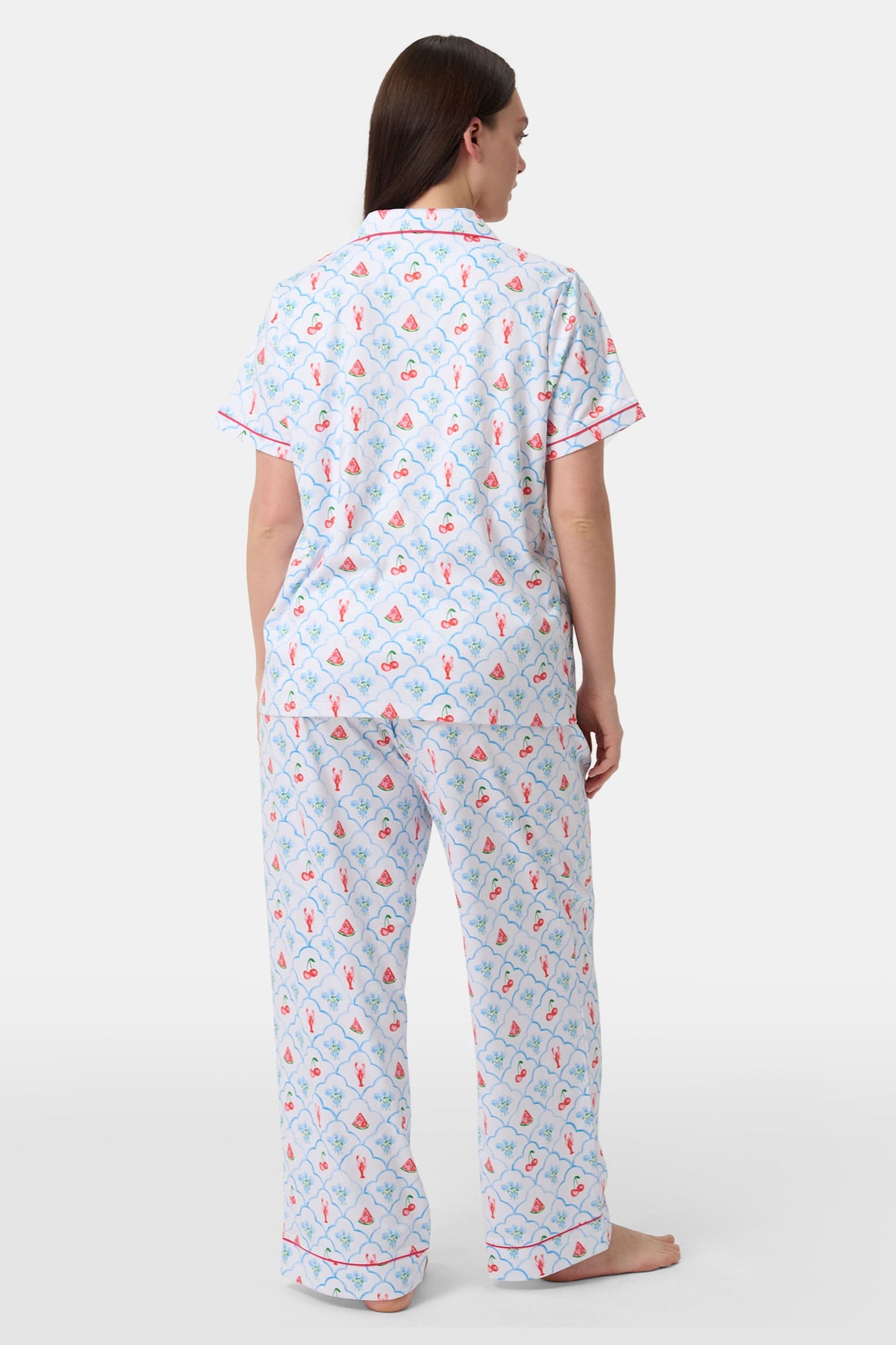 A woman in Roller Rabbit’s 4th of Jemina Fiora Pajamas and pink slippers relaxes on the bow of a blue boat at a marina while another person in white sleepwear stands nearby, partially out of frame.