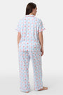A woman in Roller Rabbit’s 4th of Jemina Fiora Pajamas and pink slippers relaxes on the bow of a blue boat at a marina while another person in white sleepwear stands nearby, partially out of frame.