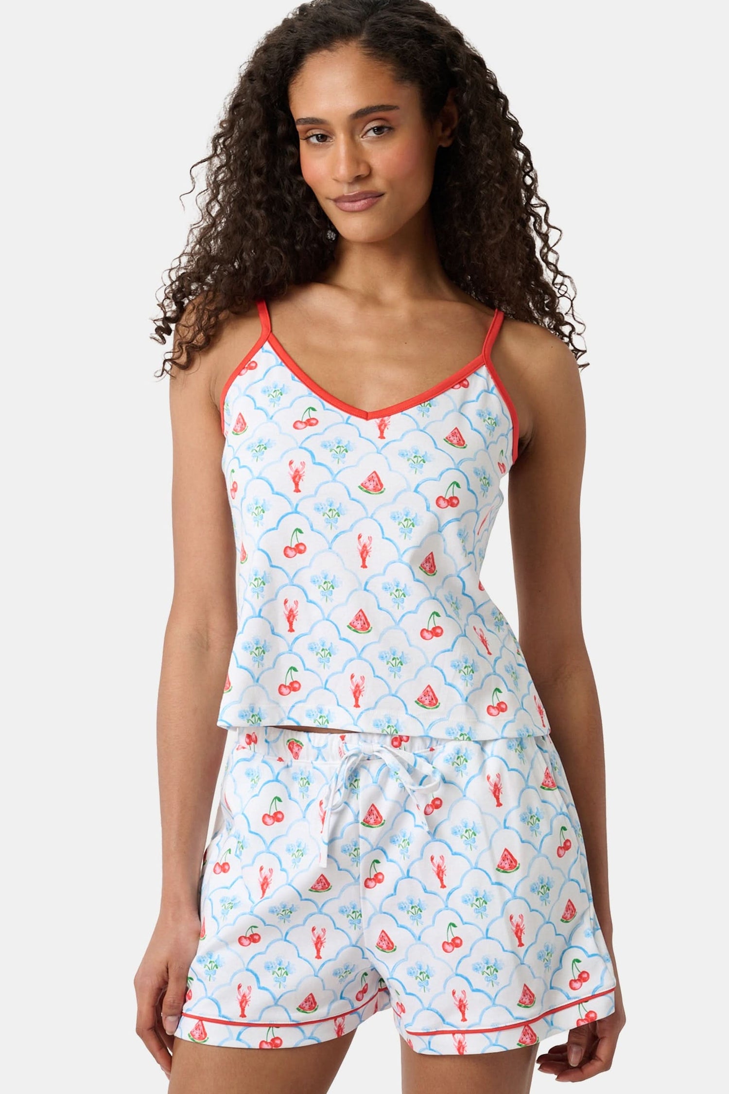 A woman with curly hair wears the Roller Rabbit 4th of Jemina Mischa Set, a Pima Cotton cami and shorts loungewear set with a white base, colorful fruit and flower prints, red trim, and drawstring shorts edged in red.