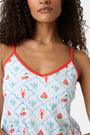 A woman with curly hair wears the Roller Rabbit 4th of Jemina Mischa Set, a Pima Cotton cami and shorts loungewear set with a white base, colorful fruit and flower prints, red trim, and drawstring shorts edged in red.