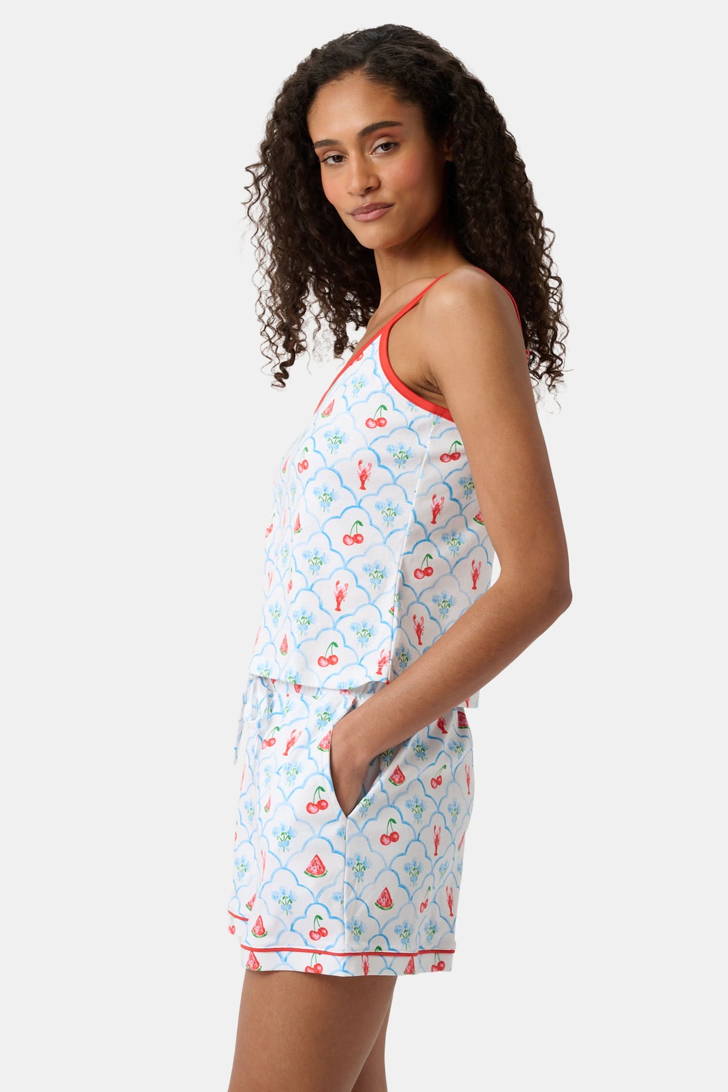 A woman with curly hair wears the Roller Rabbit 4th of Jemina Mischa Set, a Pima Cotton cami and shorts loungewear set with a white base, colorful fruit and flower prints, red trim, and drawstring shorts edged in red.