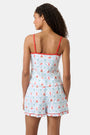 A woman with curly hair wears the Roller Rabbit 4th of Jemina Mischa Set, a Pima Cotton cami and shorts loungewear set with a white base, colorful fruit and flower prints, red trim, and drawstring shorts edged in red.