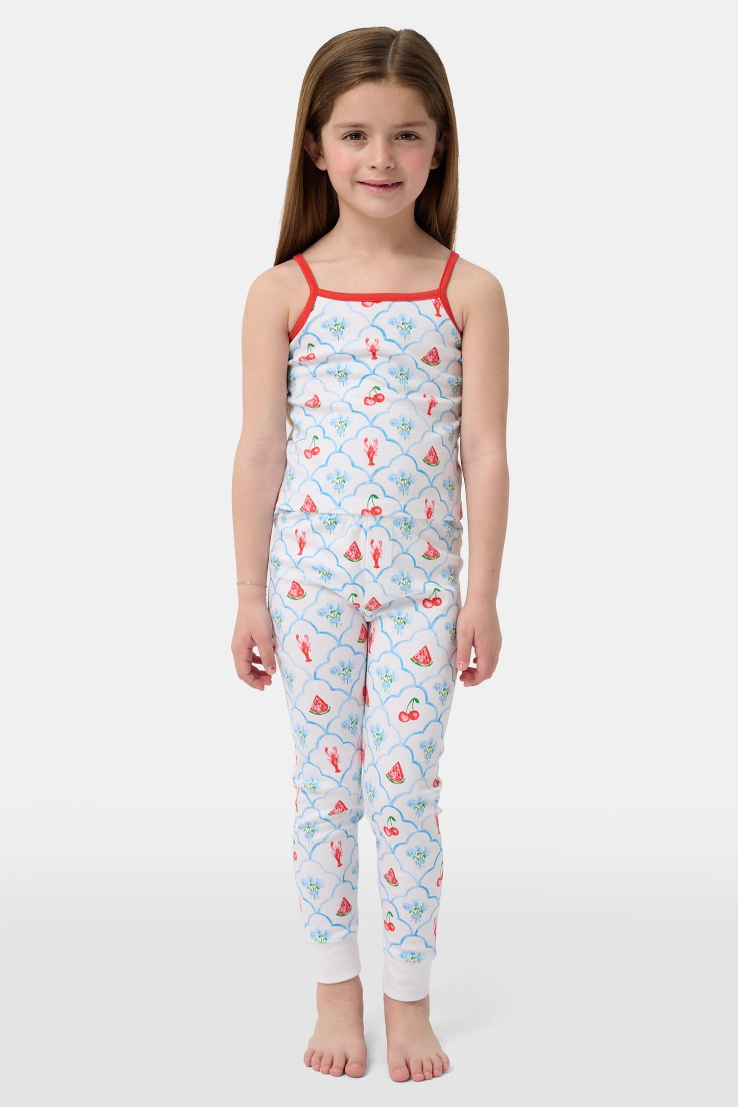 A young girl stands barefoot, smiling against a light background. She wears the Roller Rabbit Kids 4th of Jemina Noa Set—matching white Pima Cotton pajamas with red trim and a colorful flowers, butterflies, and birds print.