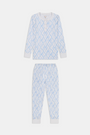 A young girl with long brown hair stands barefoot in Roller Rabbit Kids Seaside Knots Juniper Pajamas, featuring matching white and light blue knot print on soft Pima fabric with long sleeves and button details, against a plain light background.