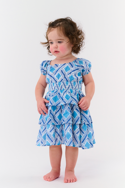 A young girl with long brown hair stands smiling against a plain light background, wearing the Kids Liana Alana Dress—a blue and white patterned cotton sundress with short sleeves, a smocked waist, and a peplum hem.