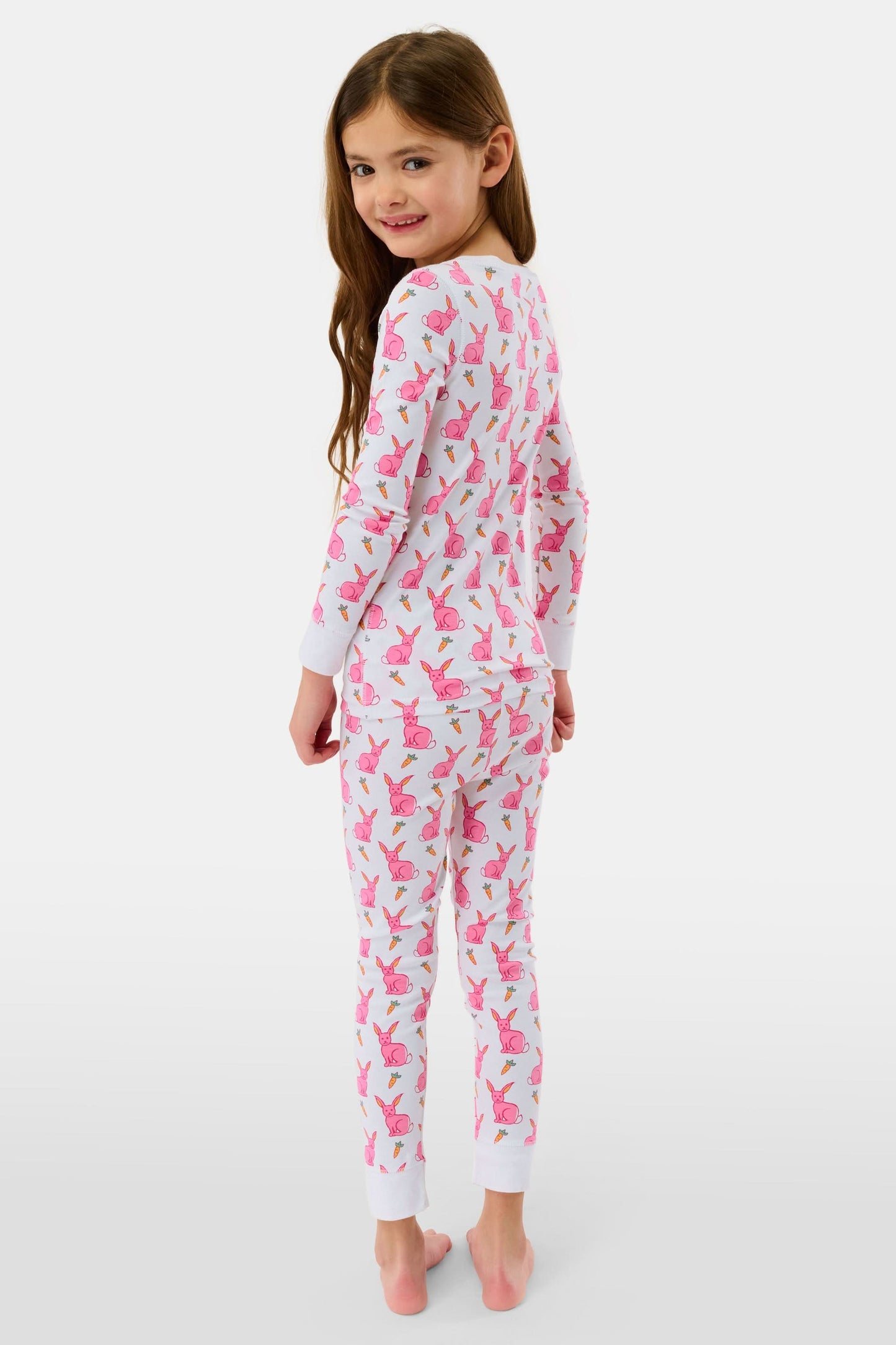 A young girl smiles barefoot in Roller Rabbit’s Kids Romy the Rabbit Pajamas, a soft Pima cotton long-sleeve set with a playful rabbit print; her brown hair frames her face against a simple, light background.