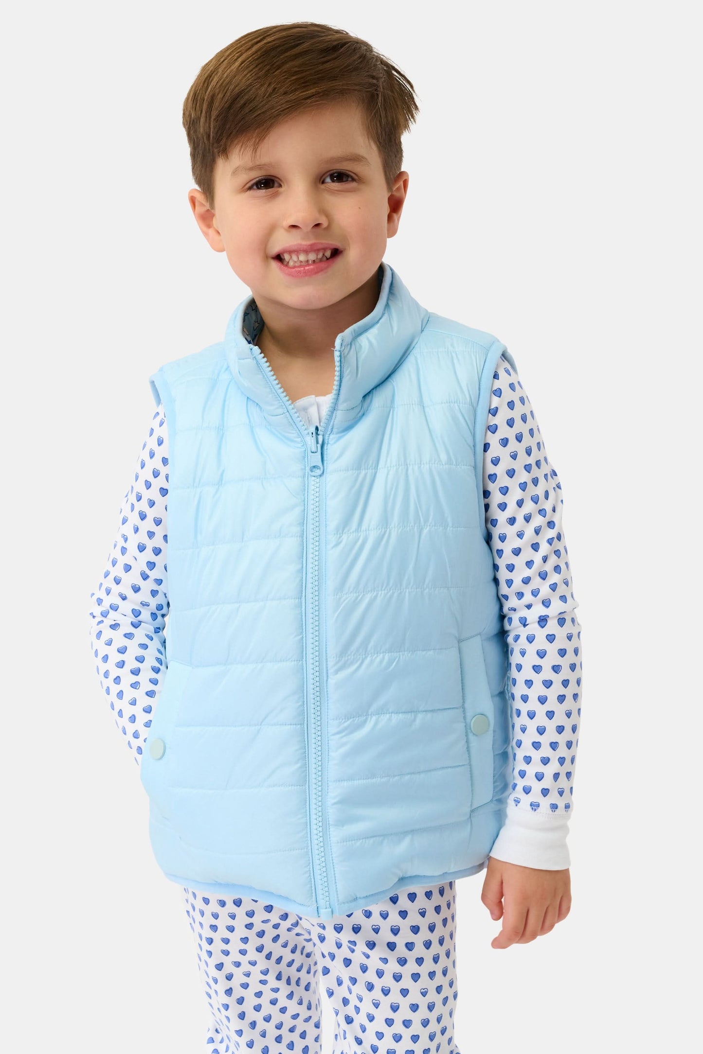 A smiling young boy wears the Kids Pop Stars Kiana Reversible Puffer Vest in sky blue over white pajamas with small blue heart patterns, standing against a plain white background.