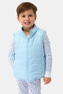 A smiling young boy wears the Kids Pop Stars Kiana Reversible Puffer Vest in sky blue over white pajamas with small blue heart patterns, standing against a plain white background.