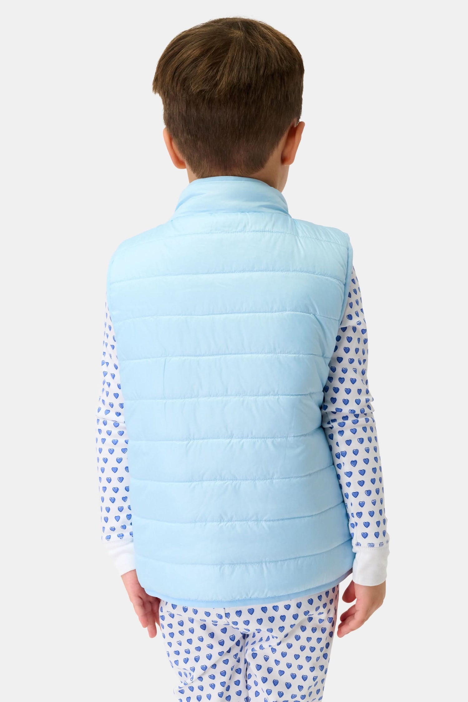 A young child with short brown hair is seen from behind, wearing the Kids Pop Stars Kiana Reversible Puffer Vest over white pajamas with a blue heart pattern, standing against a plain light background.