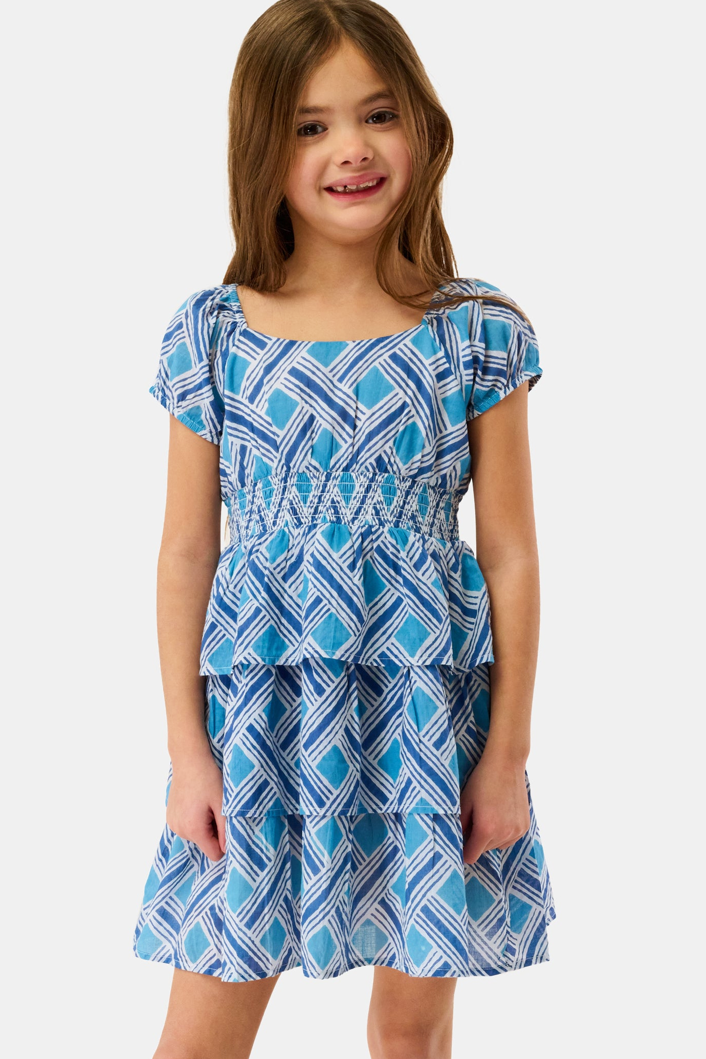 A young girl with long brown hair stands smiling against a plain light background, wearing the Kids Liana Alana Dress—a blue and white patterned cotton sundress with short sleeves, a smocked waist, and a peplum hem.