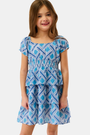 A young girl with long brown hair stands smiling against a plain light background, wearing the Kids Liana Alana Dress—a blue and white patterned cotton sundress with short sleeves, a smocked waist, and a peplum hem.