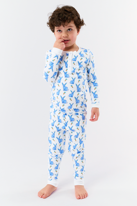 A young child with curly hair stands barefoot in white Kids Romy Rabbit Pajamas by Roller Rabbit, featuring a blue bunny print, looking at the camera and touching their mouth with one finger.