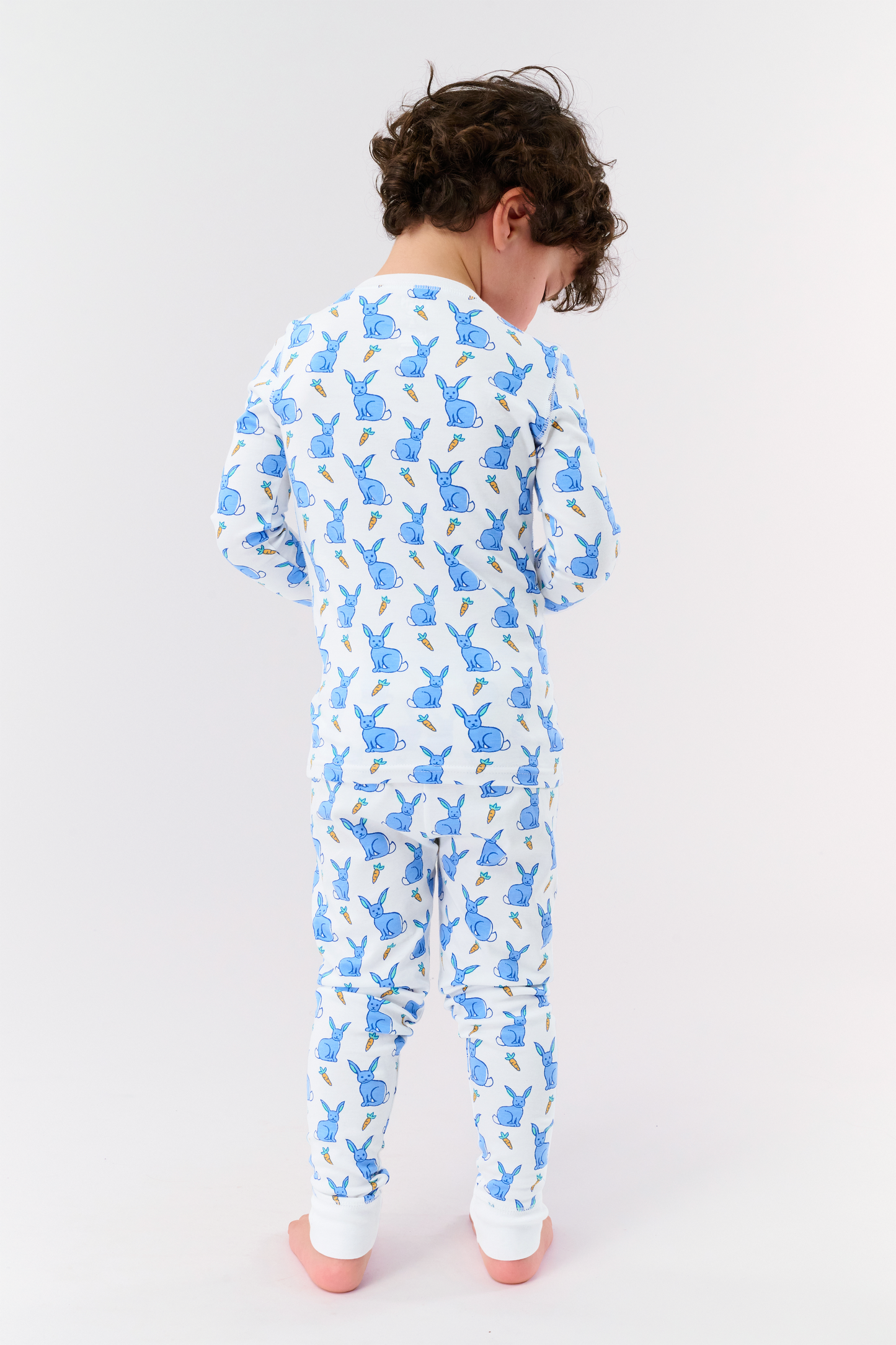 A young child with curly hair stands barefoot in white Kids Romy Rabbit Pajamas by Roller Rabbit, featuring a blue bunny print, looking at the camera and touching their mouth with one finger.