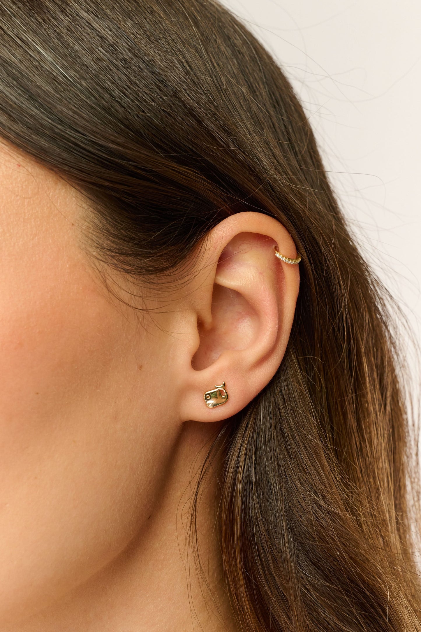 Close-up of a woman's ear with two Roller Rabbit Icon Party Earring Pack earrings: a small gold hoop on the upper cartilage and a gold and green animal stud in the lobe. Her brown hair is tucked behind her ear.