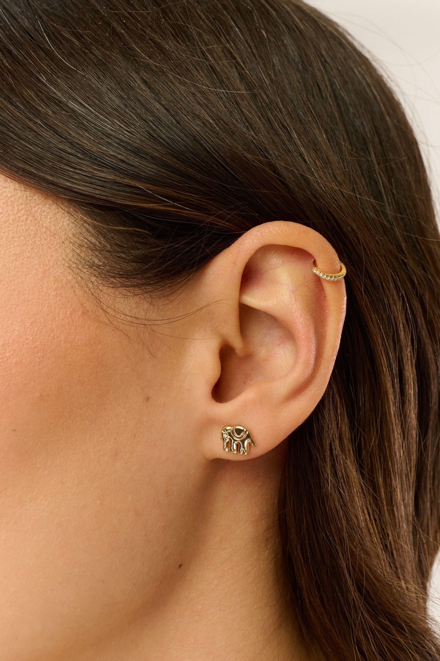 Close-up of a woman's ear with two Roller Rabbit Icon Party Earring Pack earrings: a small gold hoop on the upper cartilage and a gold and green animal stud in the lobe. Her brown hair is tucked behind her ear.