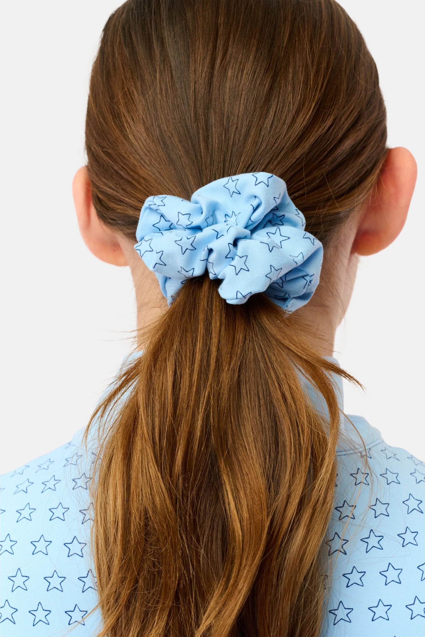 A person with long brown hair wears a star-patterned blue scrunchie from the Assorted Scrunchies Set Of 2 by Roller Rabbit, tying their hair in a low ponytail. Their light blue, star-printed shirt complements these playful accessories.