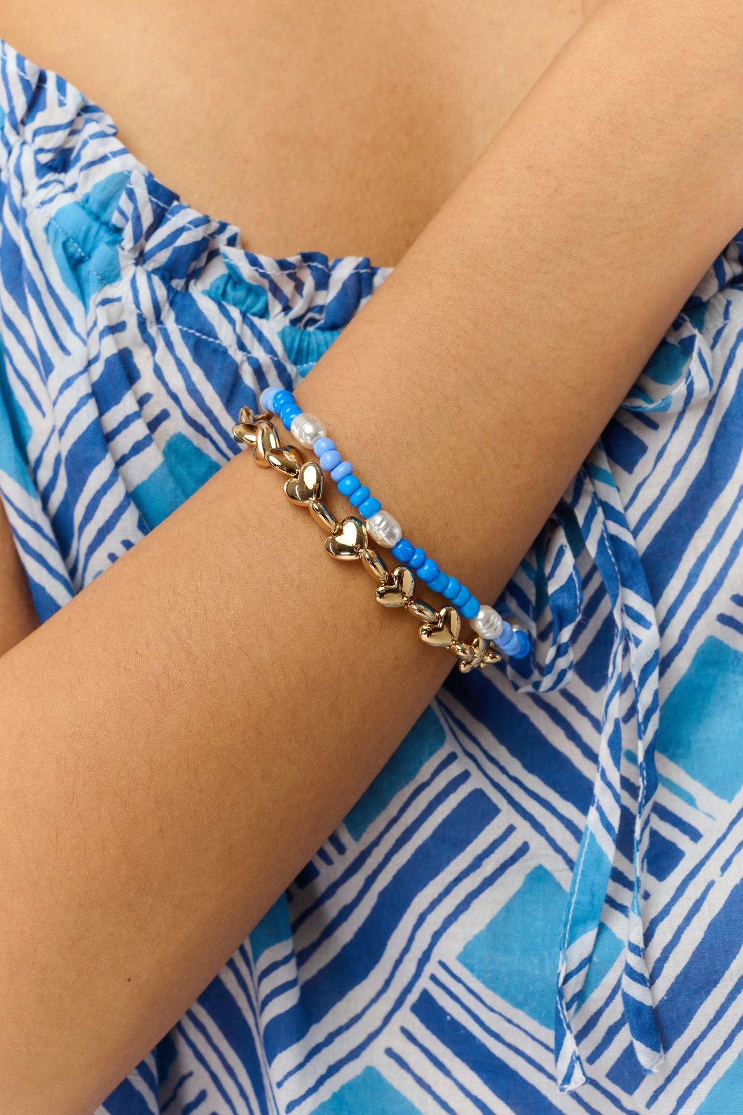 A person wearing a blue and white patterned off-shoulder top and the Roller Rabbit Colorful Hearts Bracelet Stack, featuring playful heart-shaped links and vibrant beads, on their crossed arm.