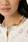 A woman is shown from the shoulders up wearing a cream knit top and the Roller Rabbit Beaded Necklace Stack, featuring colorful beaded chains and a small white flower-shaped bead.