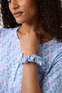 The Rosa Ditsy Monkey Scrunchie by Roller Rabbit is light blue with a playful pink monkey and floral pattern. This soft, gentle scrunchie comes in a circular shape and is displayed against a white background.