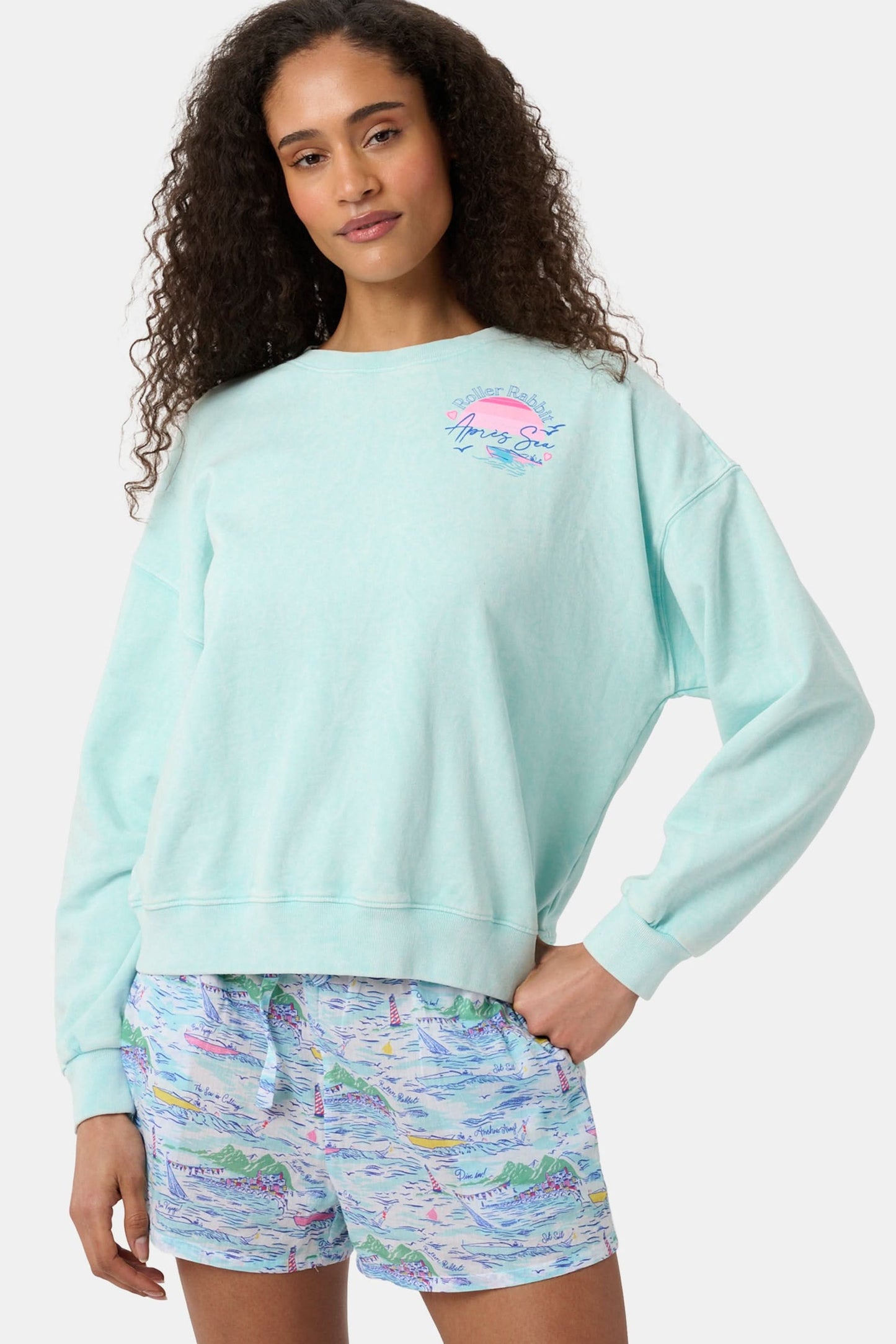 A woman wears the Roller Rabbit Apres Sea Phoebe Sweatshirt, made in the USA, mint green with a pink sunset, blue waves, birds, and "Roller Rabbit Après Sea" text on the upper left chest.