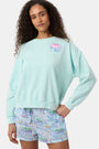 A woman wears the Roller Rabbit Apres Sea Phoebe Sweatshirt, made in the USA, mint green with a pink sunset, blue waves, birds, and 