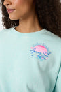 A woman wears the Roller Rabbit Apres Sea Phoebe Sweatshirt, made in the USA, mint green with a pink sunset, blue waves, birds, and 