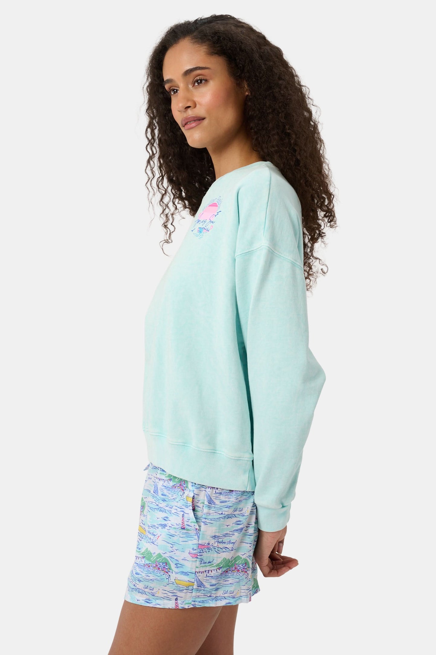 A woman wears the Roller Rabbit Apres Sea Phoebe Sweatshirt, made in the USA, mint green with a pink sunset, blue waves, birds, and "Roller Rabbit Après Sea" text on the upper left chest.