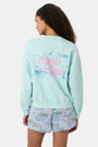 A woman wears the Roller Rabbit Apres Sea Phoebe Sweatshirt, made in the USA, mint green with a pink sunset, blue waves, birds, and 