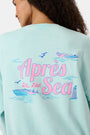 A woman wears the Roller Rabbit Apres Sea Phoebe Sweatshirt, made in the USA, mint green with a pink sunset, blue waves, birds, and 