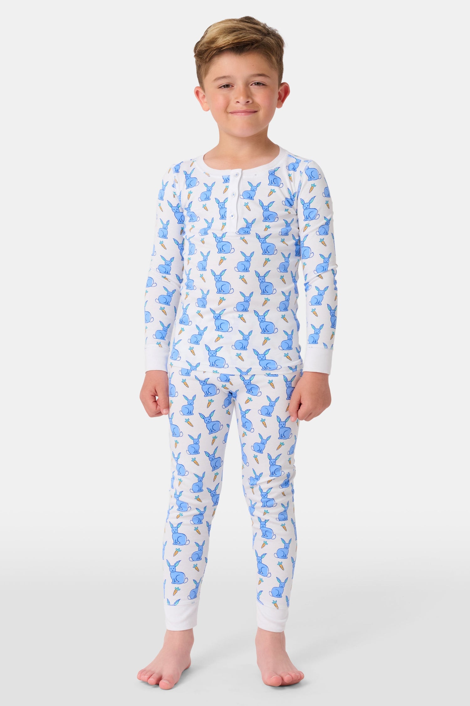 A young child with curly hair stands barefoot in white Kids Romy Rabbit Pajamas by Roller Rabbit, featuring a blue bunny print, looking at the camera and touching their mouth with one finger.