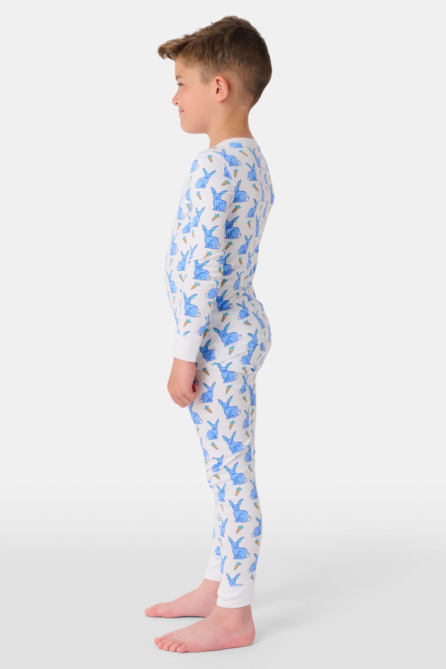 A young child with curly hair stands barefoot in white Kids Romy Rabbit Pajamas by Roller Rabbit, featuring a blue bunny print, looking at the camera and touching their mouth with one finger.