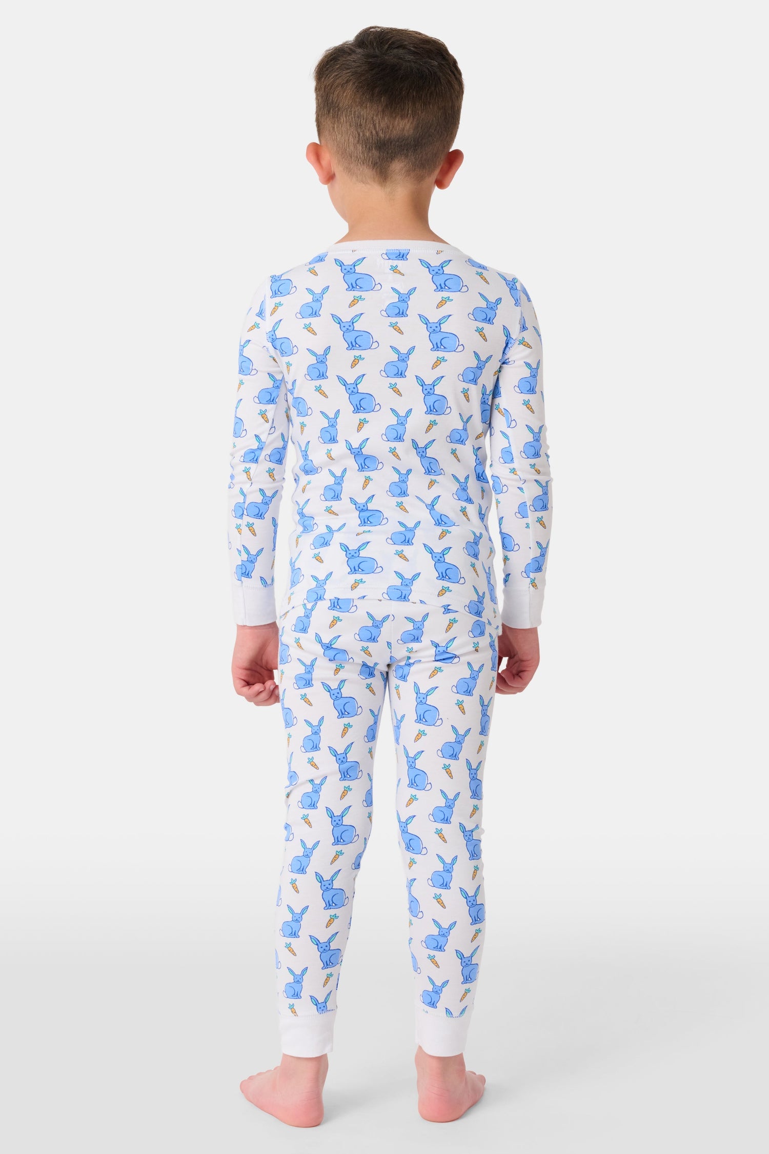 A young child with curly hair stands barefoot in white Kids Romy Rabbit Pajamas by Roller Rabbit, featuring a blue bunny print, looking at the camera and touching their mouth with one finger.