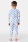 A young child with curly hair stands barefoot in white Kids Romy Rabbit Pajamas by Roller Rabbit, featuring a blue bunny print, looking at the camera and touching their mouth with one finger.