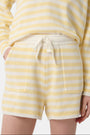 A person wears Roller Rabbit’s Striped Brie Short—a knit yellow and white horizontal striped short with a white drawstring at the elastic waistband and hands tucked in the front pockets.