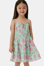 A young girl with long brown hair smiles, wearing the Roller Rabbit Kids Camellia Block Karla Dress, a sleeveless mint green cotton dress with pink floral patterns. She stands with one hand by her side and the other holding her dress.
