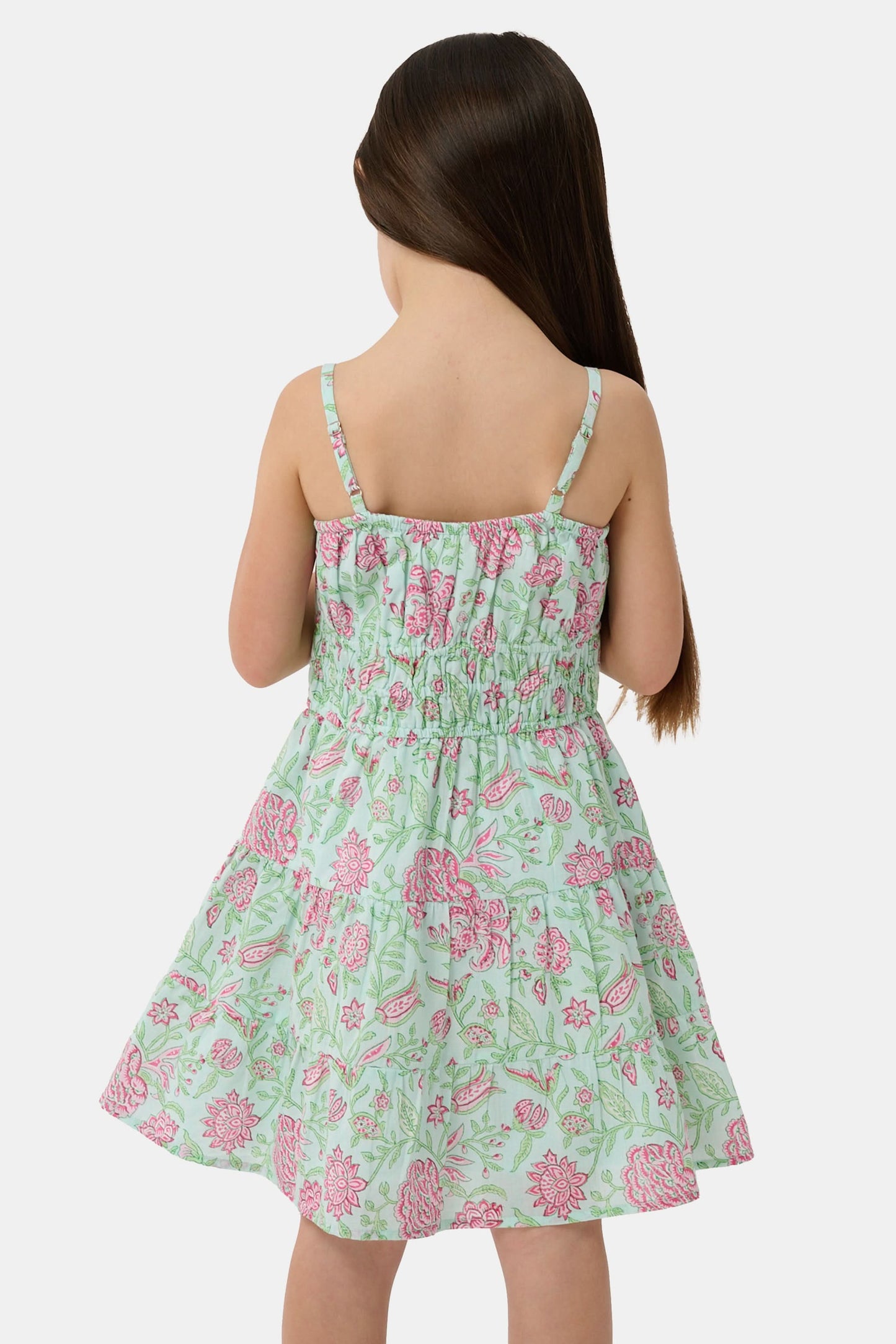 A young girl with long brown hair smiles, wearing the Roller Rabbit Kids Camellia Block Karla Dress, a sleeveless mint green cotton dress with pink floral patterns. She stands with one hand by her side and the other holding her dress.