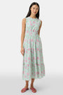 A woman with long dark hair and a neutral expression poses against a plain light background wearing the Roller Rabbit Camellia Block Milani Dress, a pastel green and pink floral cotton maxi, paired with pink sandals.