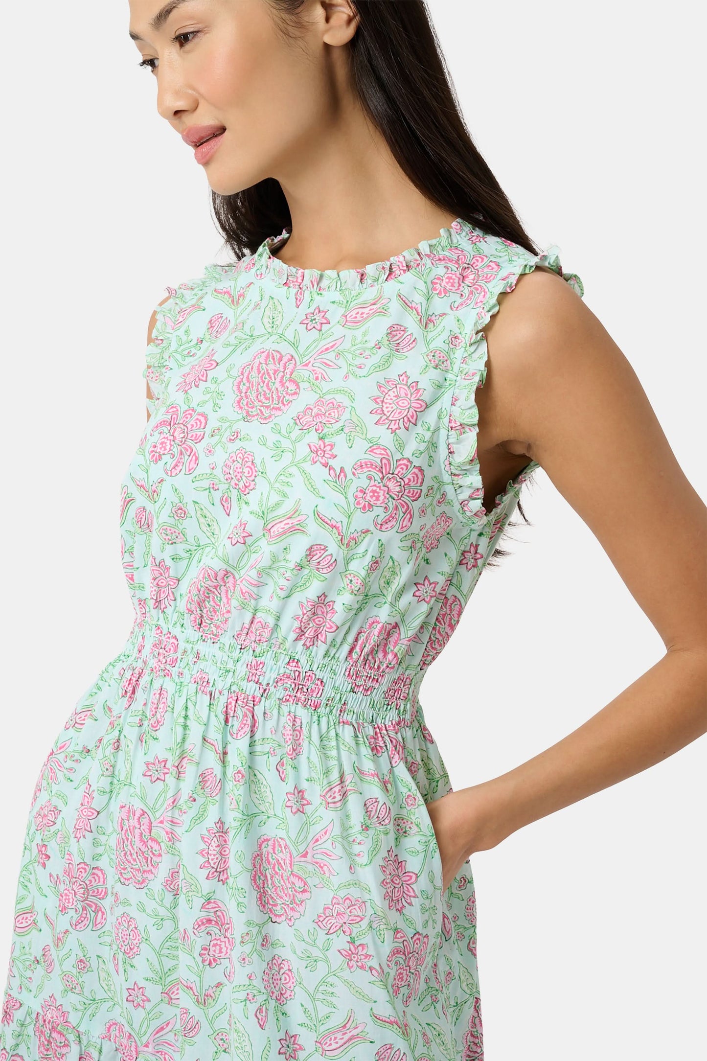 A woman with long dark hair and a neutral expression poses against a plain light background wearing the Roller Rabbit Camellia Block Milani Dress, a pastel green and pink floral cotton maxi, paired with pink sandals.