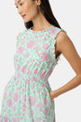 A woman with long dark hair and a neutral expression poses against a plain light background wearing the Roller Rabbit Camellia Block Milani Dress, a pastel green and pink floral cotton maxi, paired with pink sandals.