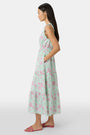 A woman with long dark hair and a neutral expression poses against a plain light background wearing the Roller Rabbit Camellia Block Milani Dress, a pastel green and pink floral cotton maxi, paired with pink sandals.