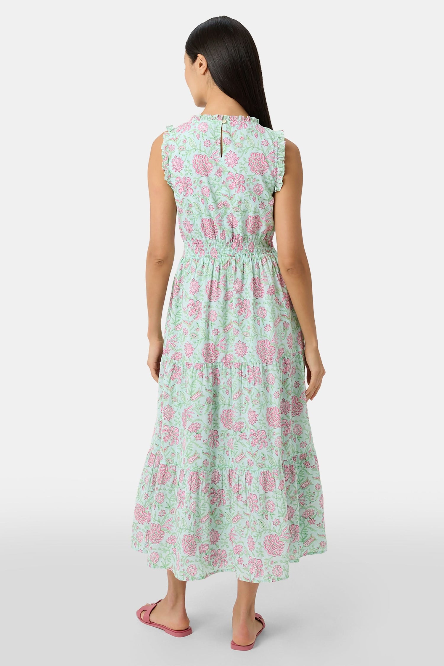A woman with long dark hair and a neutral expression poses against a plain light background wearing the Roller Rabbit Camellia Block Milani Dress, a pastel green and pink floral cotton maxi, paired with pink sandals.
