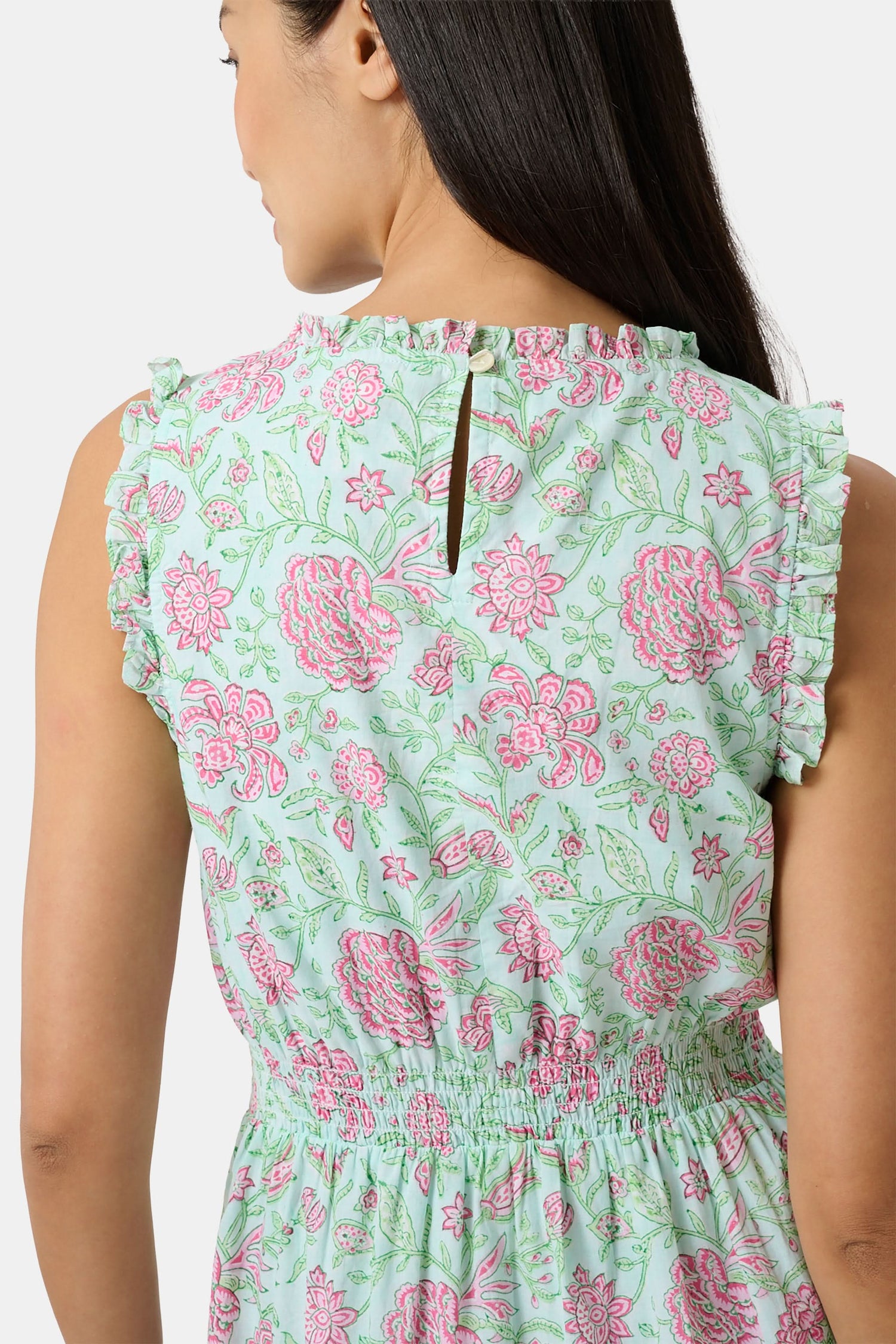 A woman with long dark hair and a neutral expression poses against a plain light background wearing the Roller Rabbit Camellia Block Milani Dress, a pastel green and pink floral cotton maxi, paired with pink sandals.